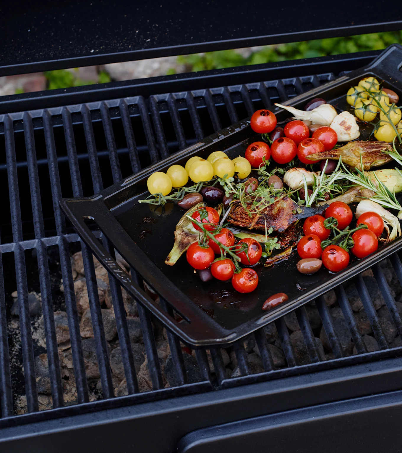 A black grilling pan filled with roasted vegetables including red and yellow cherry tomatoes, garlic, artichoke, and olives, on a grill.