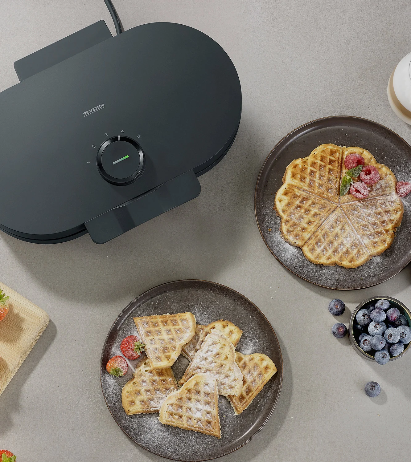 Dark grey waffle maker next to two plates of heart-shaped waffles with berries.
