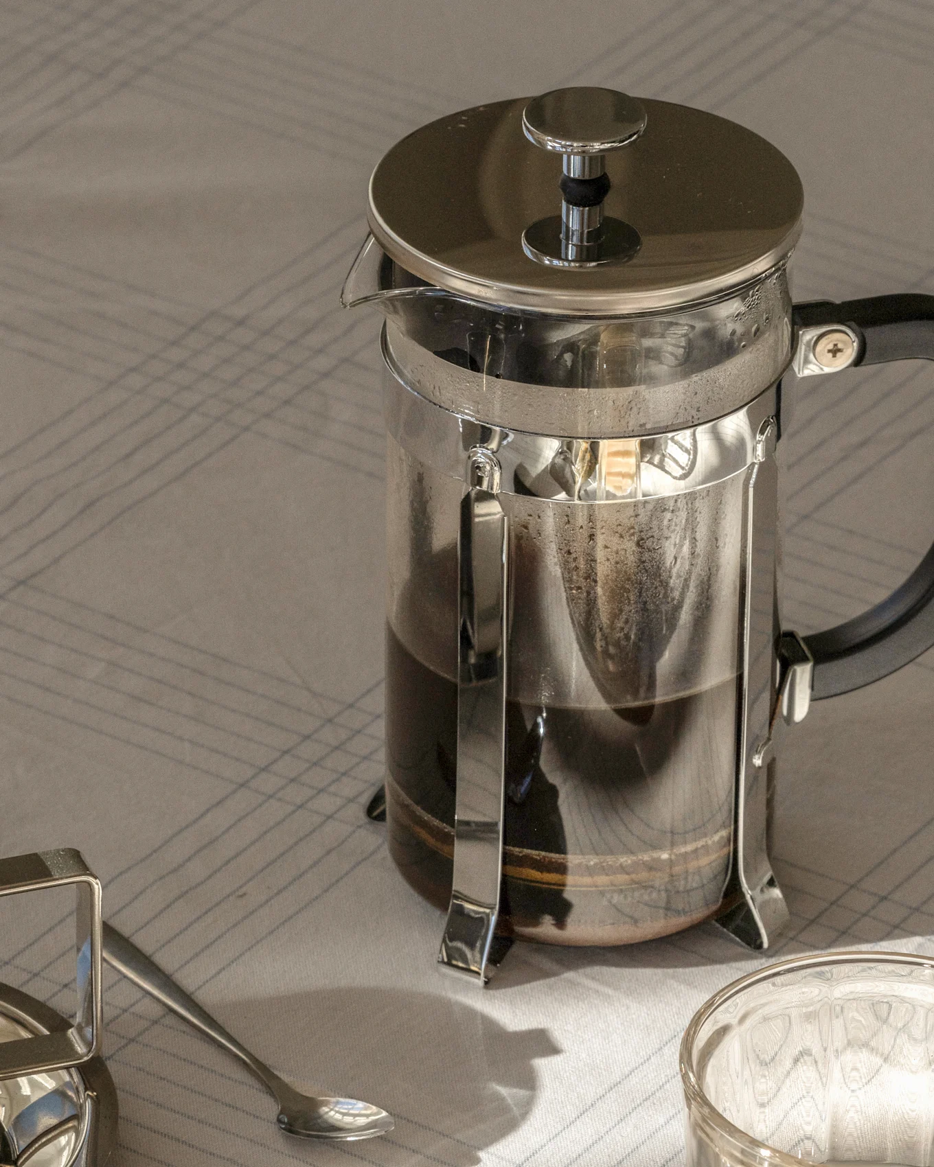 French press coffee maker with dark coffee on a striped tablecloth, next to a spoon and glass.