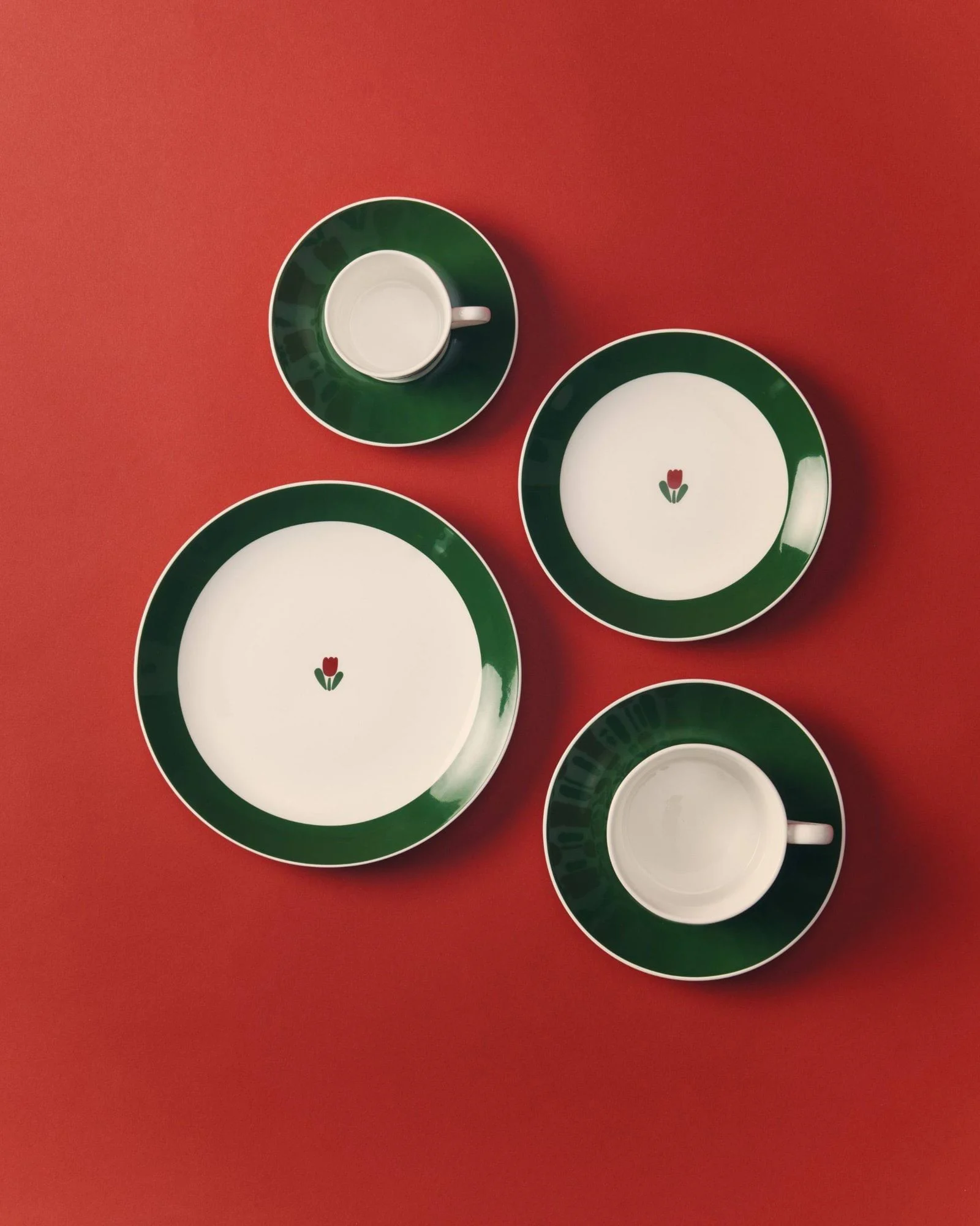 Four white and green tableware items on a red background: two plates with a red tulip design and two cups with saucers.
