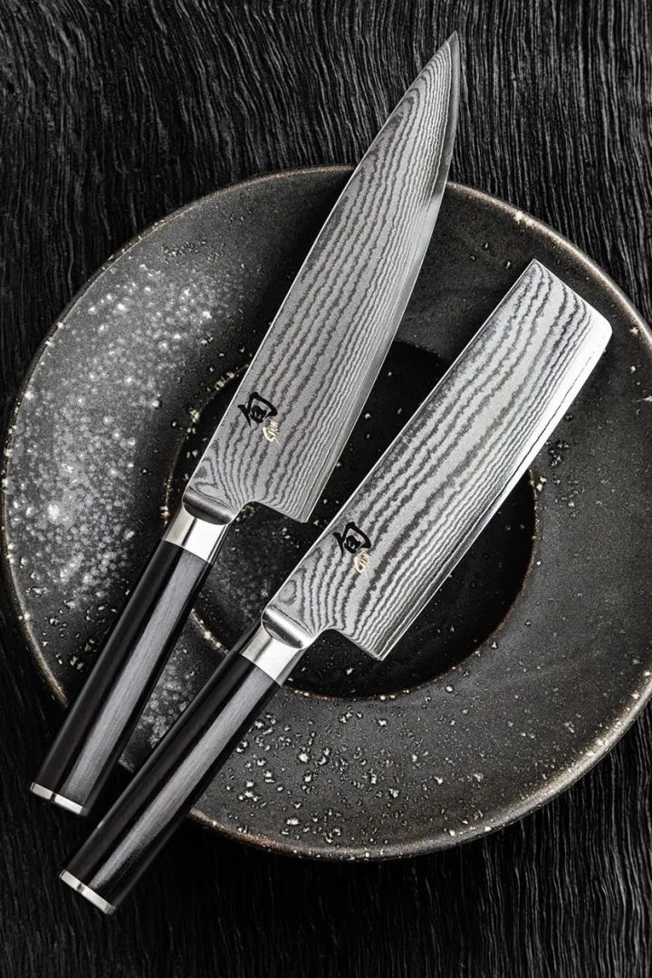 A pair of Kai Shun Japanese knives, a gyuto and a nakiri with black handles are laid on top of a black plate. 