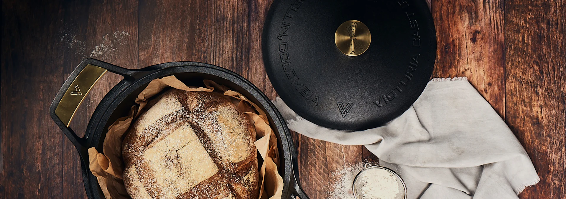 Freshly baked, scored bread in a black cast iron Dutch oven with gold handles, its lid, and a sieve of flour on a rustic wooden table.