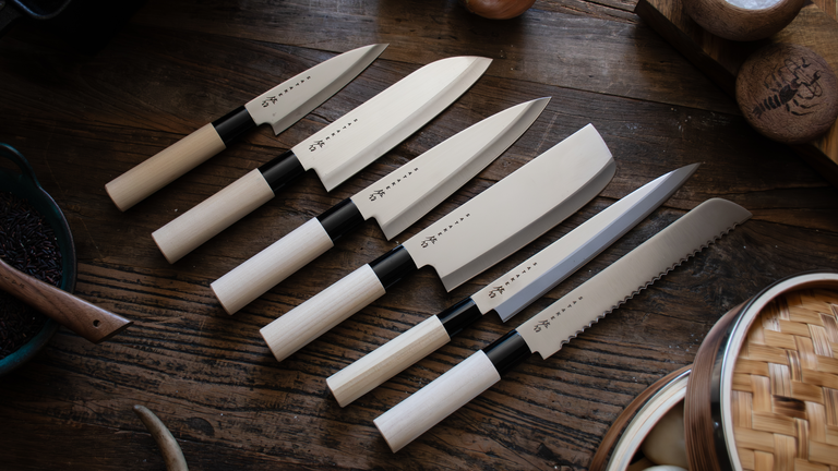 A collection of various different Japanese knives laid out in a row on a wooden kitchen sideboard. 