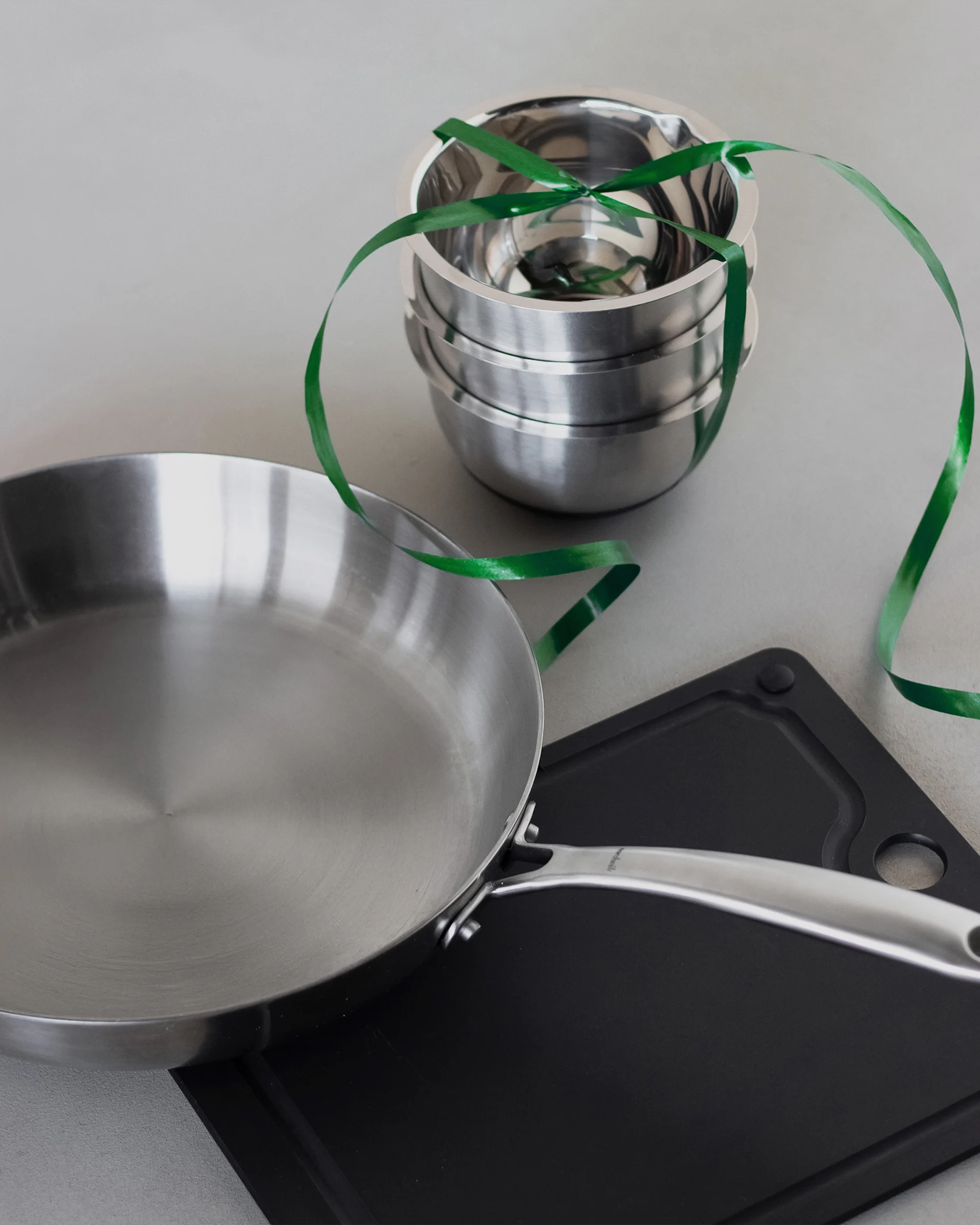 A stainless steel frying pan, a stack of metal mixing bowls tied with a green ribbon, and a black cutting board on a light grey surface.