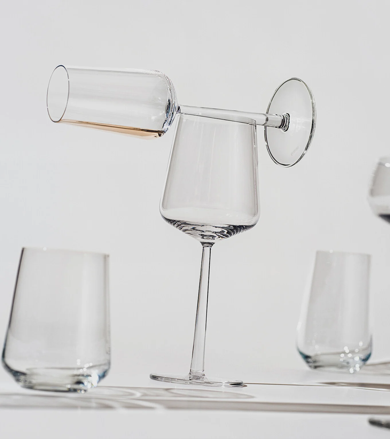 A champagne flute with amber liquid balances horizontally on the rim of an upright wine glass, with other clear glasses in the background.