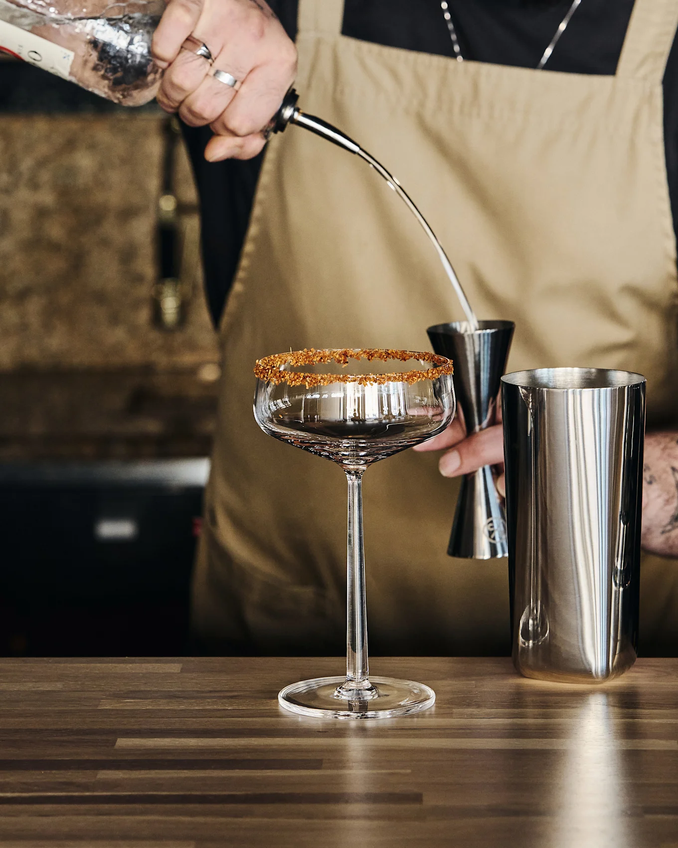 Bartender pouring liquid from a bottle into a jigger over a mixing tin, next to a stemmed glass with a sugared rim.
