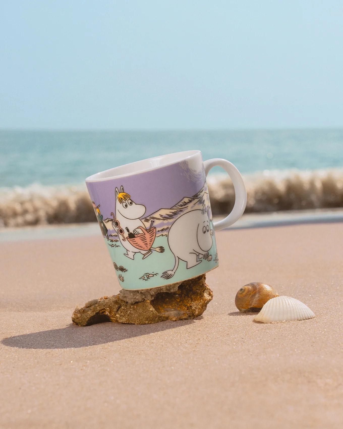 Moomin mug on beach, featuring Snorkmaiden and Moomintroll, tilted on a rock with seashells and ocean waves.