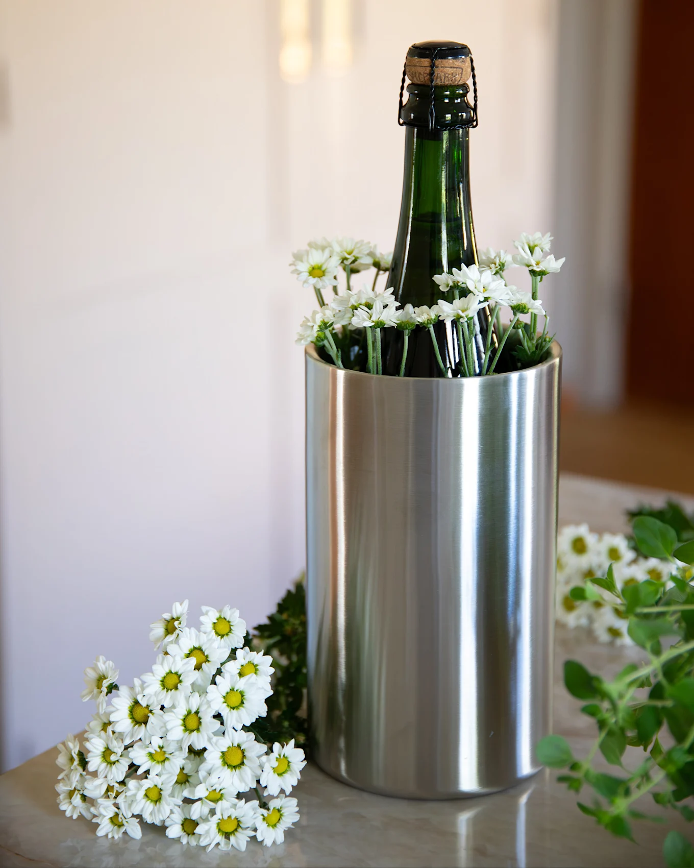 A dark green bottle with a cork stopper in a stainless steel cooler, surrounded by white daisy flowers on a light marble surface.