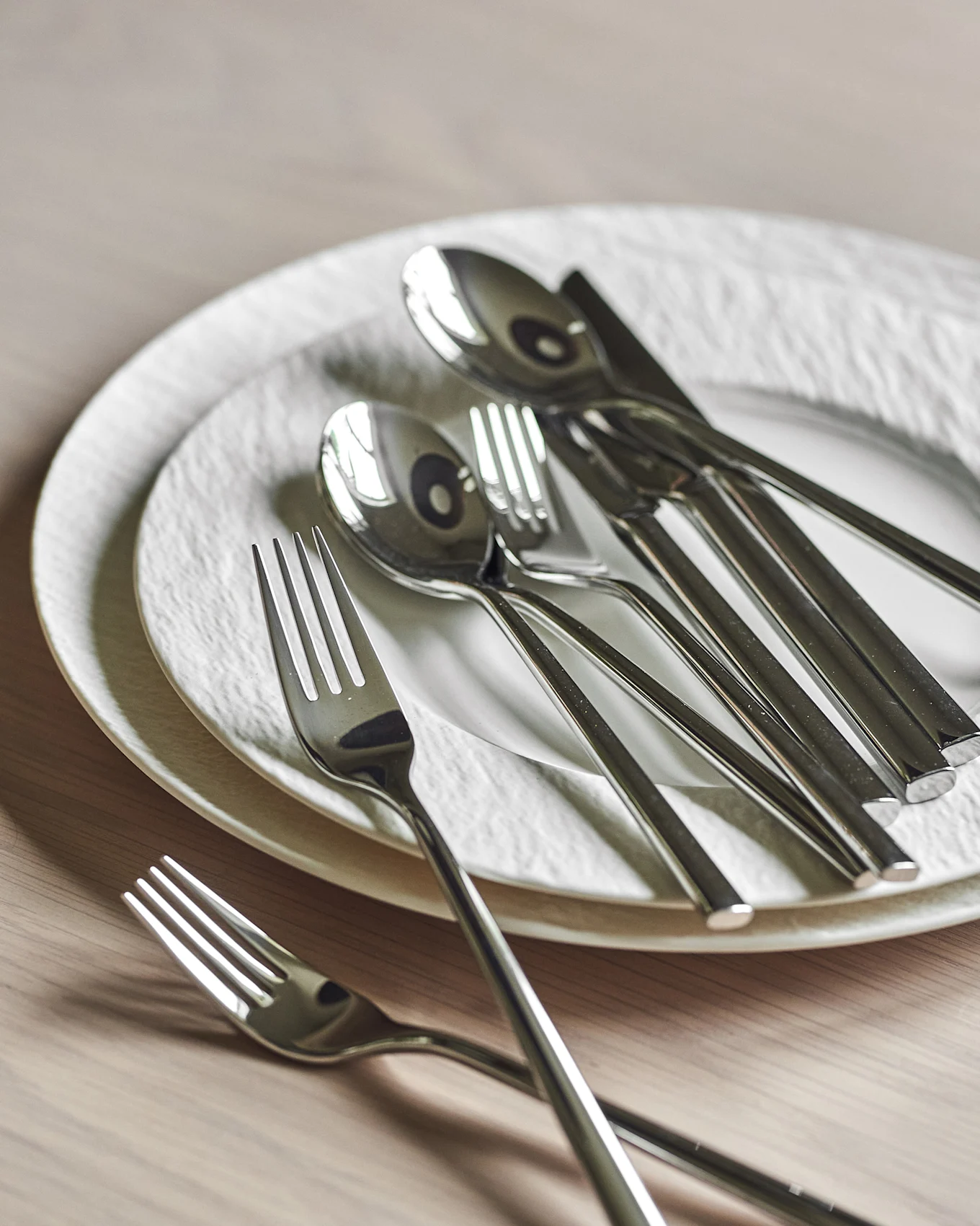 A stack of two white textured plates with a collection of sleek, shiny silver cutlery arranged on top and beside them on a wooden table.
