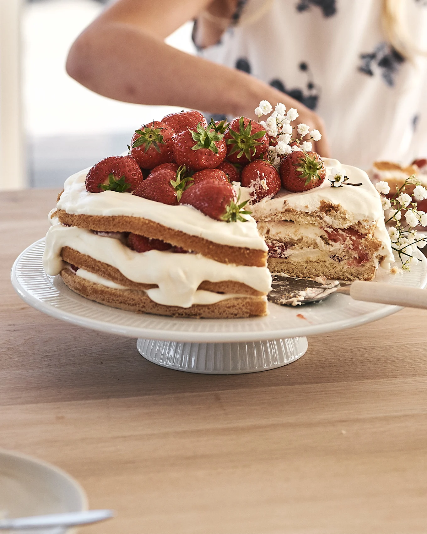 A strawberry layer cake with whipped cream and fresh strawberries on a white fluted cake stand. A slice has been cut.