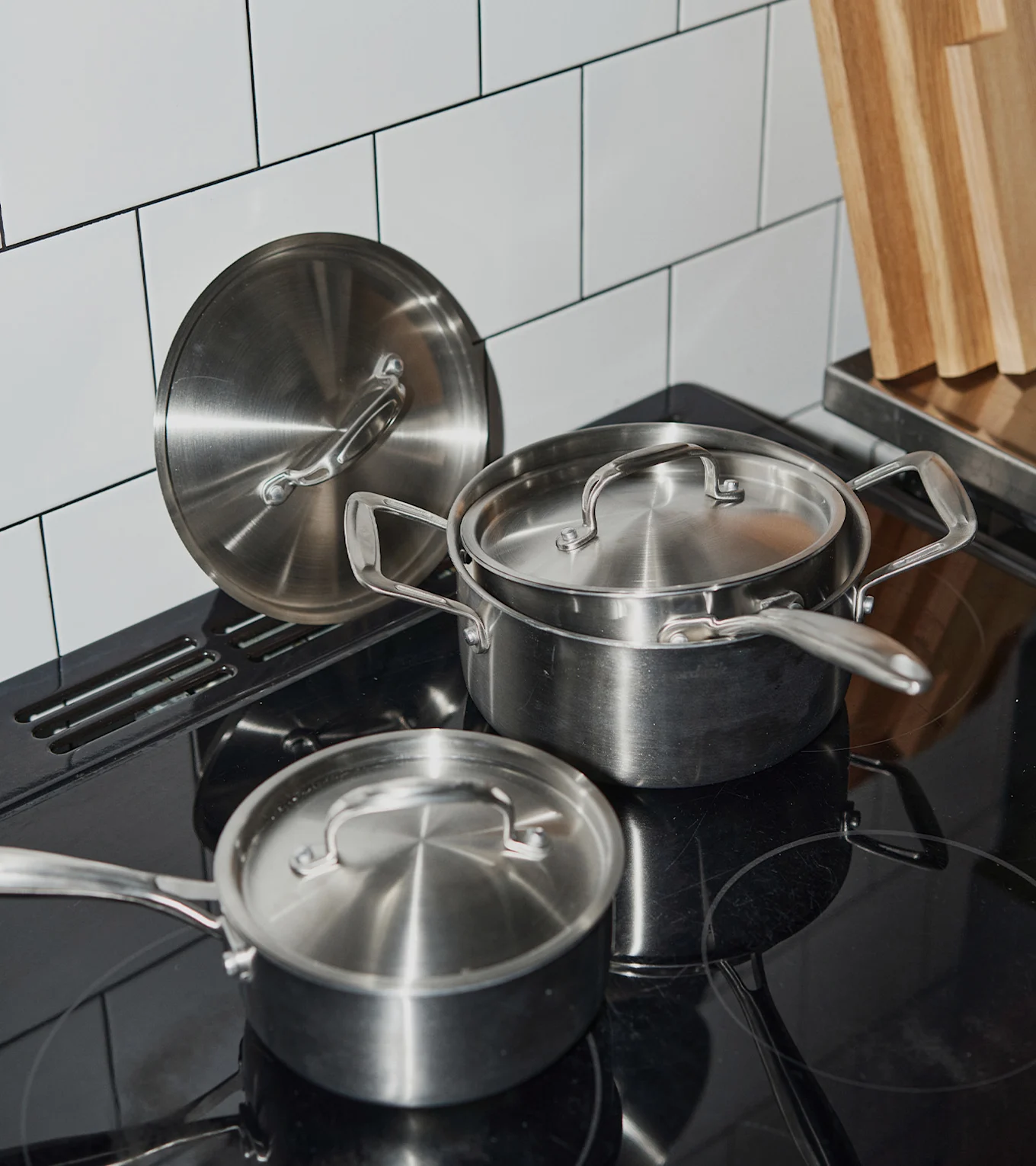 Stainless steel pots and lids on a black induction stovetop, with white tiled backsplash and wooden cutting boards.