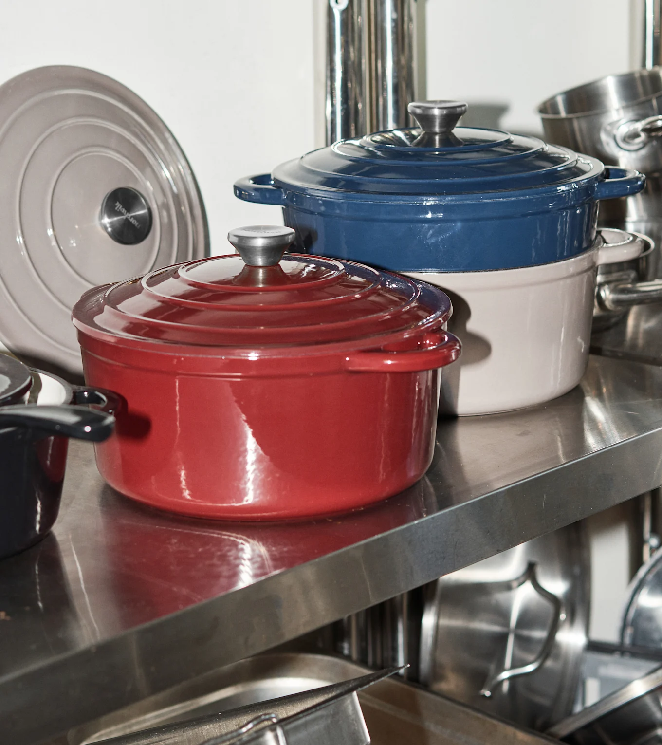 Three enameled cast iron pots in red, blue, and light beige on a metal shelf with a gray pot lid.