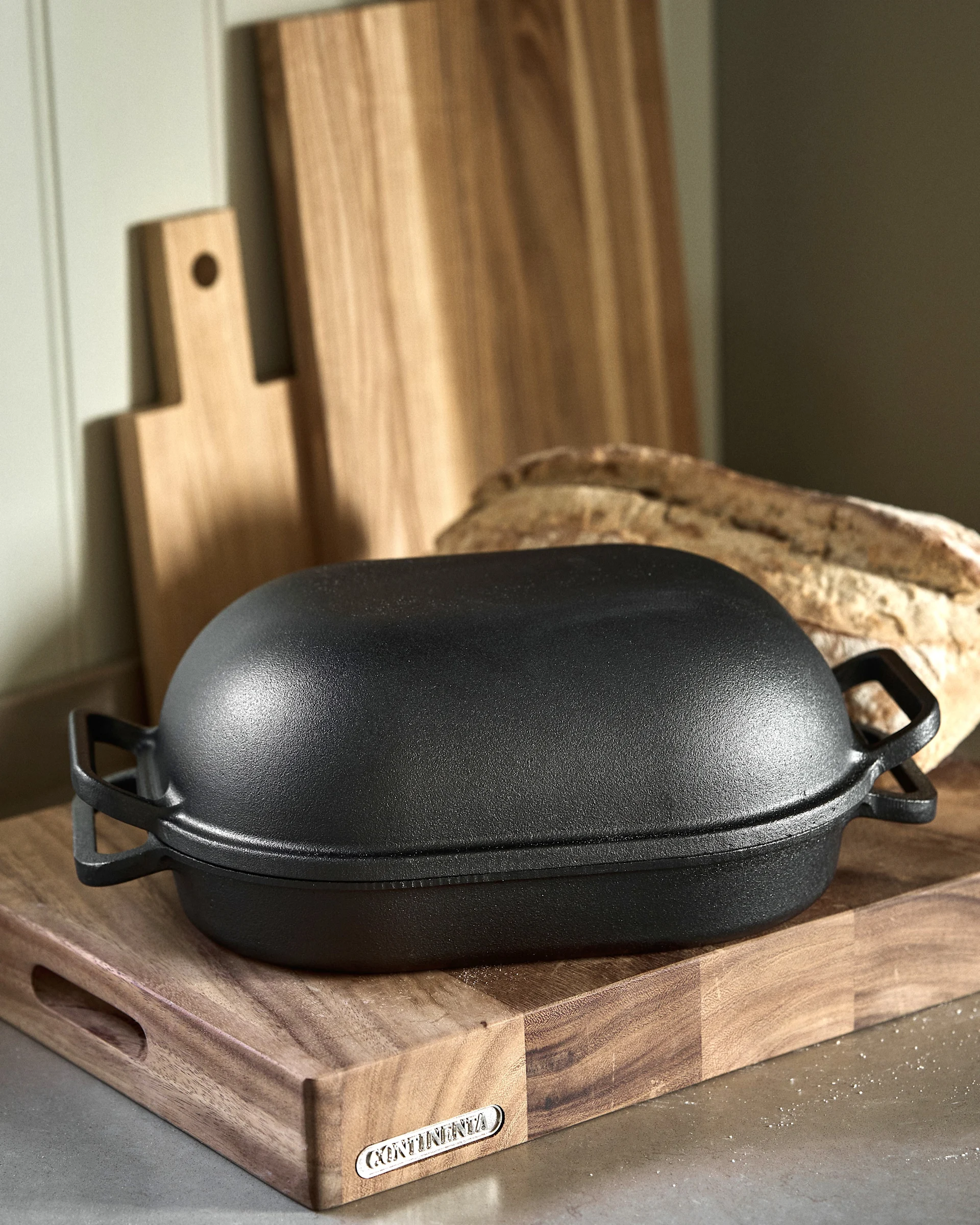 Black oval cast iron baker on a wooden cutting board with a baguette and other boards in the background. The cutting board has a 'Continenta' brand tag.