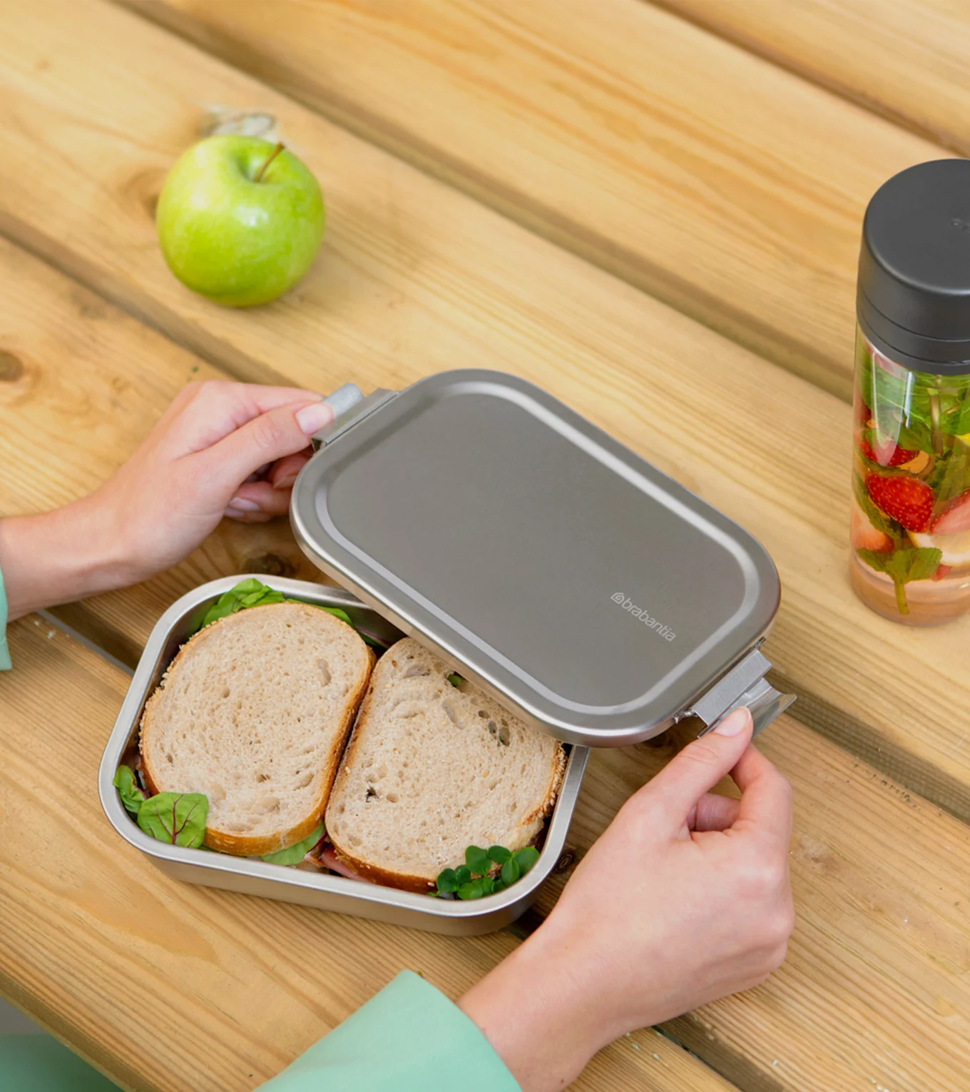 Hands opening a silver lunchbox with sandwiches, an apple, and a fruit-infused water bottle on a wooden table.