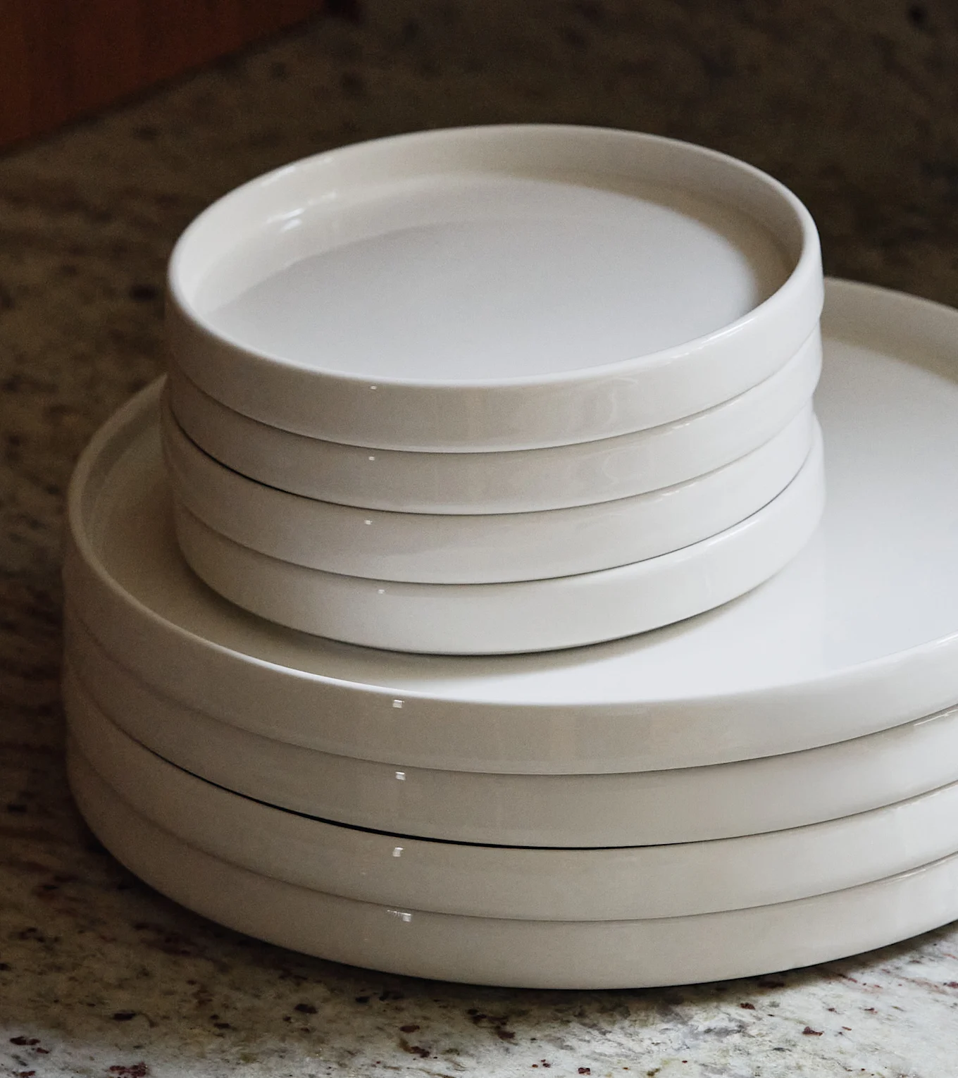 Two stacks of white ceramic plates and shallow bowls on a speckled countertop.
